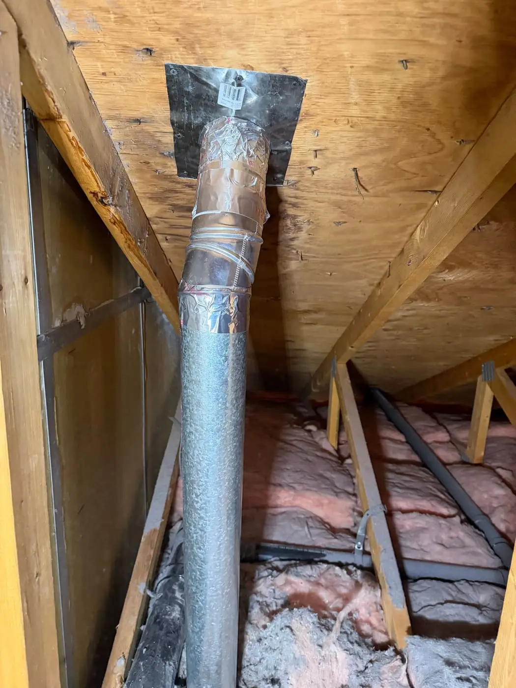 A rigid metal vent pipe with foil tape on its joints, rising through insulation to the roof deck of an attic.
