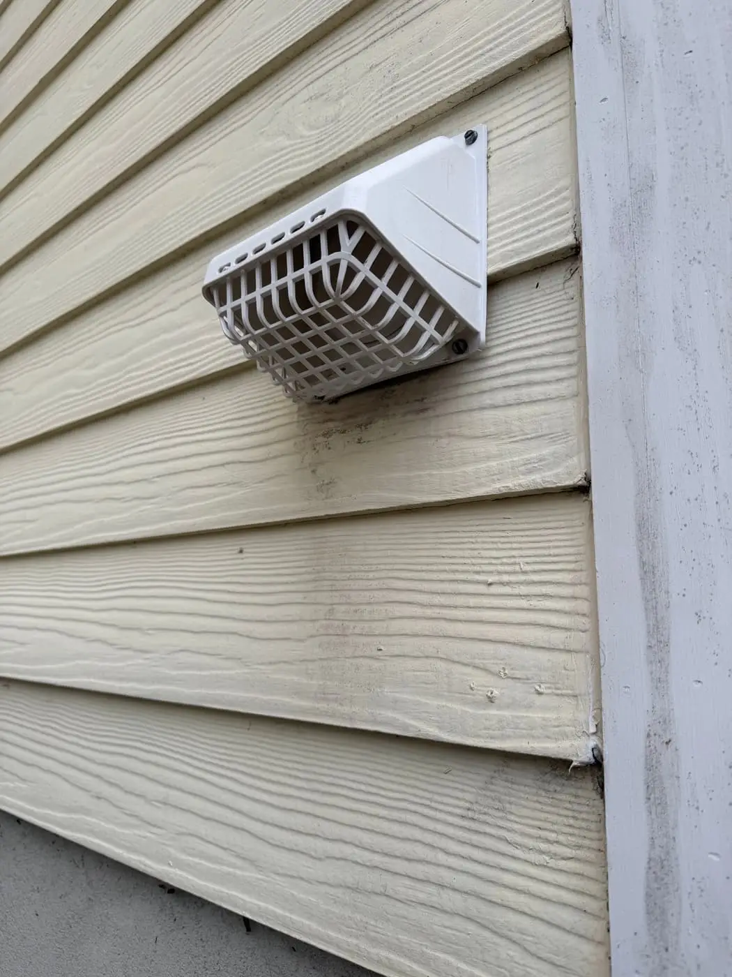 A white dryer vent hood with a guard grille, mounted on beige siding.