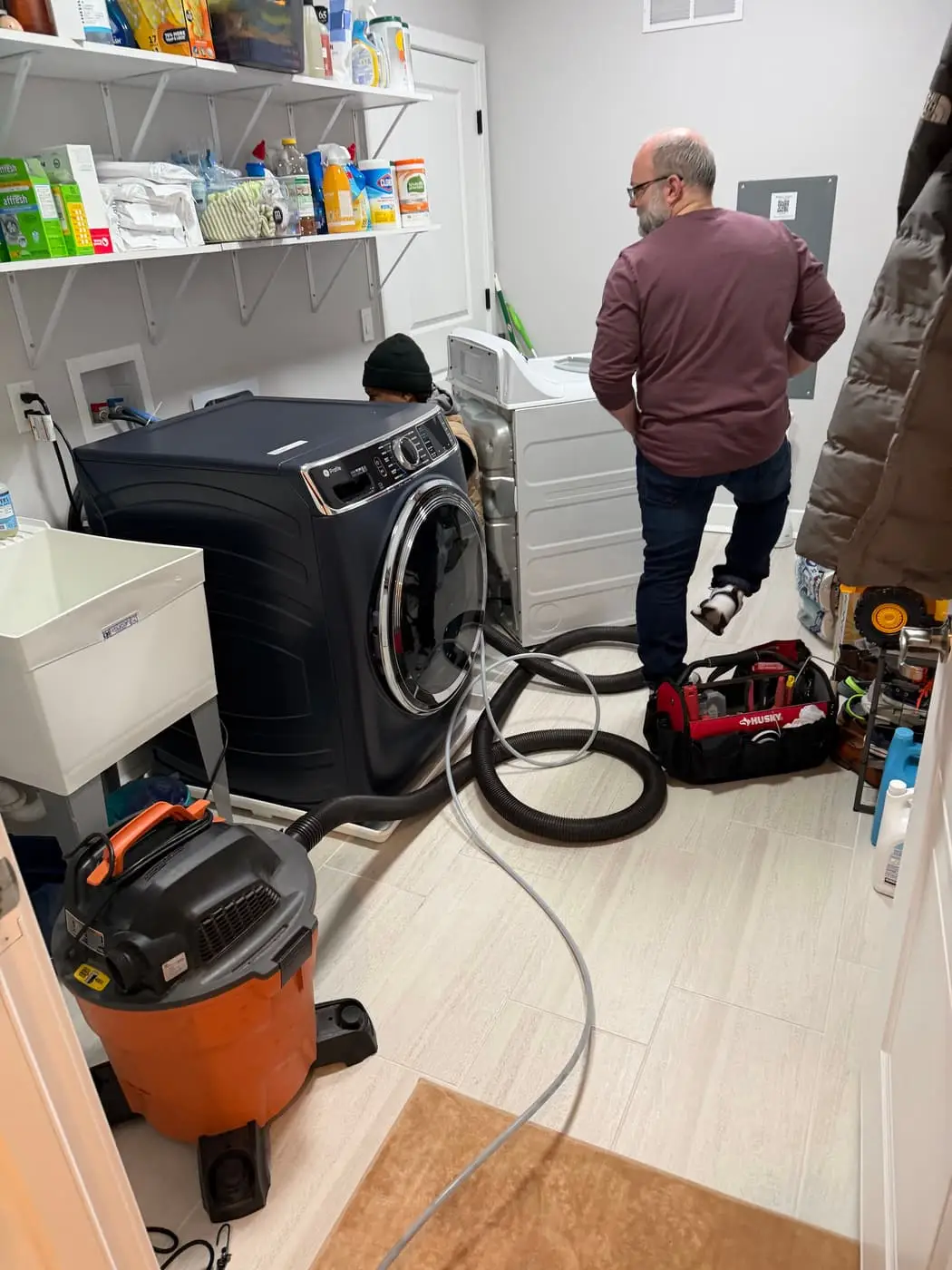 A laundry room with the dryer pulled away from the wall, a shop vacuum and hoses on the floor, and two people standing by the machines.