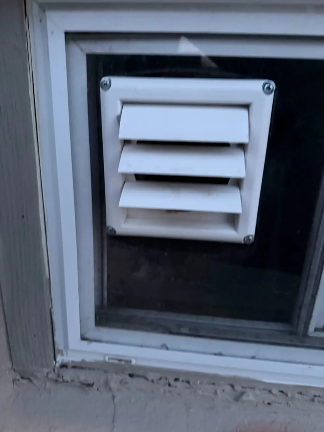 A white louvered vent hood set into a panel that fills a basement window.